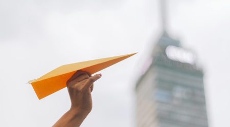 Papierflieger orange in Hand vor Hochhaus Papierflieger wird in die Luft geworfen und von einer Hand festgehalten, im Hintergrund ein Hochhaus und der Himmel
