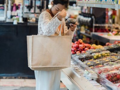 Frau mit Einkaufstasche Junge Frau mit Stofftasche prüft ein Produkt in der Obst- und Gemüseabteilung eines modernen Supermarkts